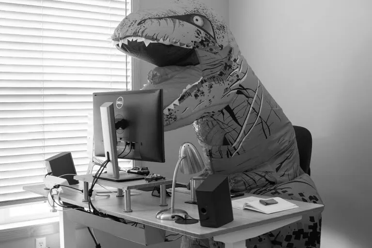 A person in a t-rex costume sitting at a computer desk